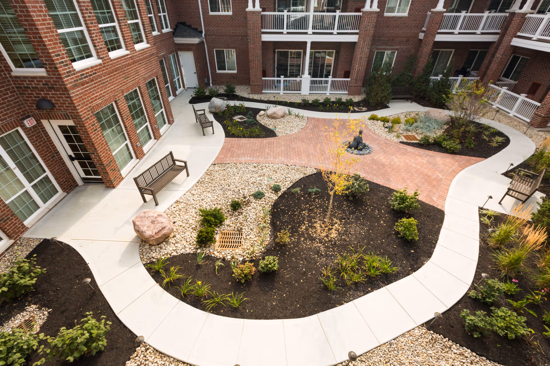 View of a landscaped courtyard in a senior living facility with brick buildings surrounding it. The courtyard features curved concrete walkways, planted garden beds with small shrubs and a young tree, decorative rocks, benches, and a small water fountain in the center.