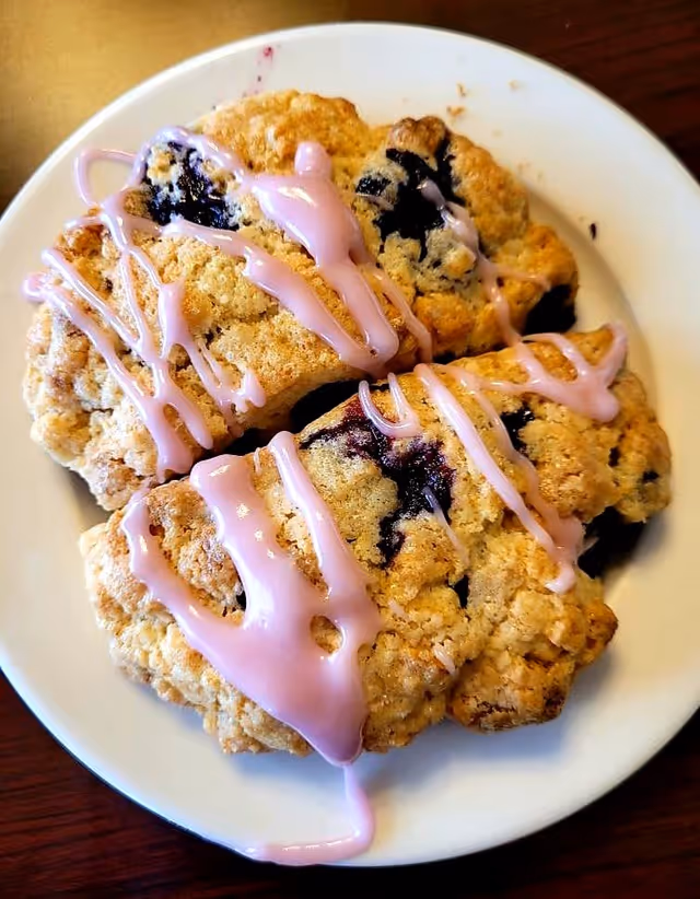 A white plate with three blueberry scones drizzled with pink icing on a wooden table.