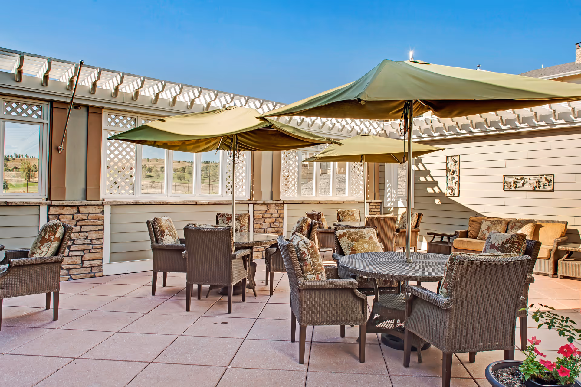 Outdoor patio area with multiple round tables, each shaded by large green umbrellas. The tables are surrounded by wicker chairs with floral cushions. The patio is enclosed by walls with lattice windows and stone accents, and there are some potted plants visible.