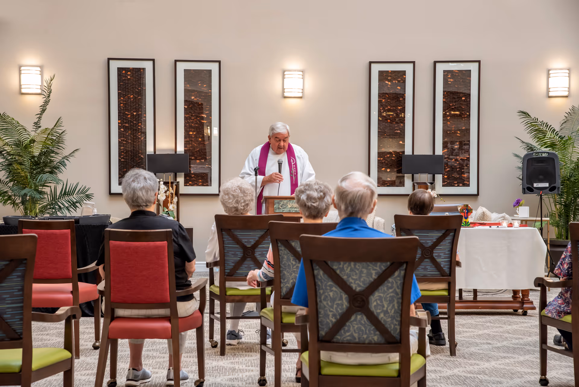 An elderly group seated in chairs facing a man in religious attire speaking at a podium in a well-lit room with framed artwork and plants on the walls.