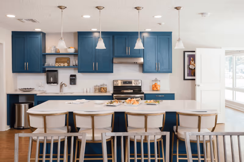 Modern kitchen with blue cabinets, white countertops, and a central island with four white cushioned bar stools. The kitchen features a stainless steel stove and oven, three pendant lights hanging above the island, and various kitchen items such as jars, bowls, and a fruit container on the counters. There is a window and a door on the right side, and a framed artwork on the wall.