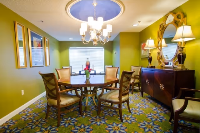 A bright dining room with a round wooden table surrounded by six chairs, a chandelier overhead, a sideboard with lamps and mirror, and a patterned carpet.