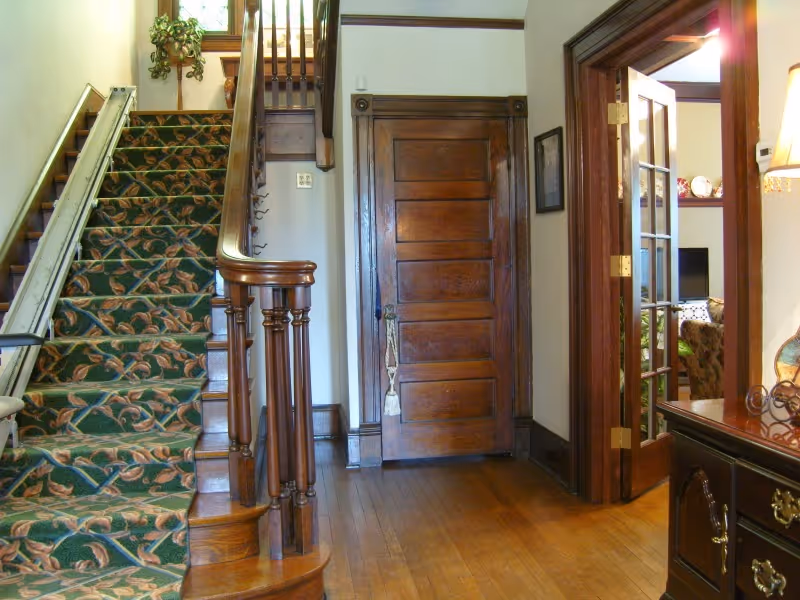 Interior view of a hallway in Fitzwater's Assisted Living featuring a wooden staircase with a floral carpet runner and a stair lift on the left. There is a closed wooden door straight ahead and an open doorway with glass-paneled double doors on the right, leading to a living room area with a television and furniture. The floor is wooden, and there is a small plant on a stand near the top of the stairs.
