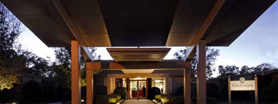 Front entrance of Bedford Care Center of Hattiesburg at dusk with a covered porte-cochere, illuminated walkway, and a sign.