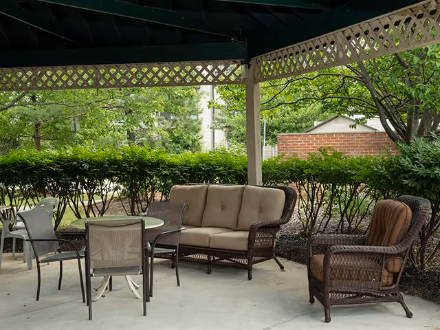 Outdoor covered seating area with a cushioned wicker loveseat, a cushioned wicker armchair, and a glass-top table surrounded by four chairs. The area is surrounded by green bushes and trees, with a brick wall and a small shed visible in the background.