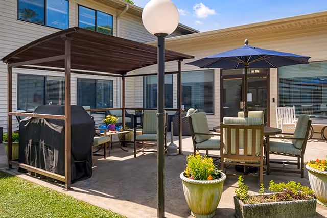Outdoor patio area at Brookdale Hendersonville East featuring a covered grill, multiple seating arrangements with cushioned chairs, a table with a large blue umbrella, potted plants, and a lamp post, all set against the backdrop of the facility building with windows.
