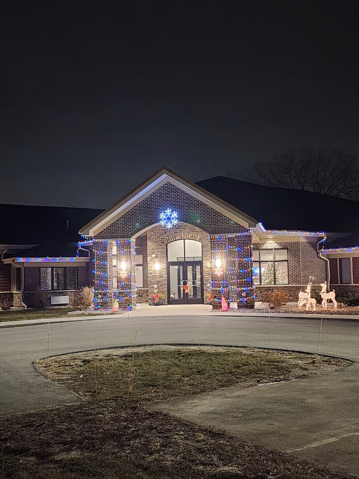Night view of the front entrance of CopperStone Assisted Living decorated with colorful Christmas lights, a large illuminated snowflake above the door, and lighted reindeer figures on the right side. The building is made of brick with large windows and a circular driveway in front.