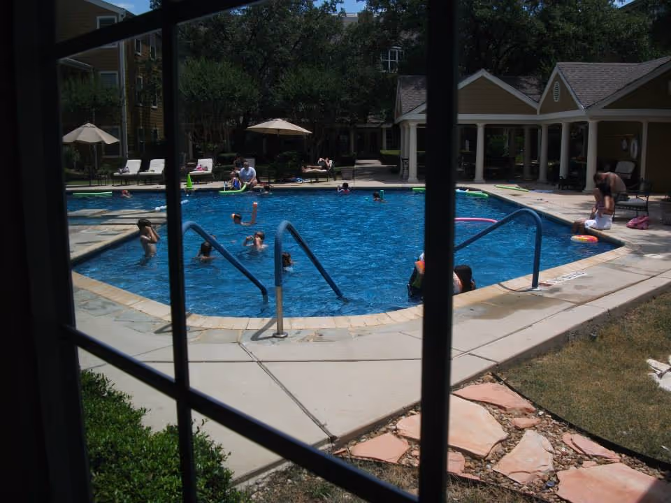 View through window bars of a community outdoor swimming pool with people swimming and lounging on the surrounding deck.
