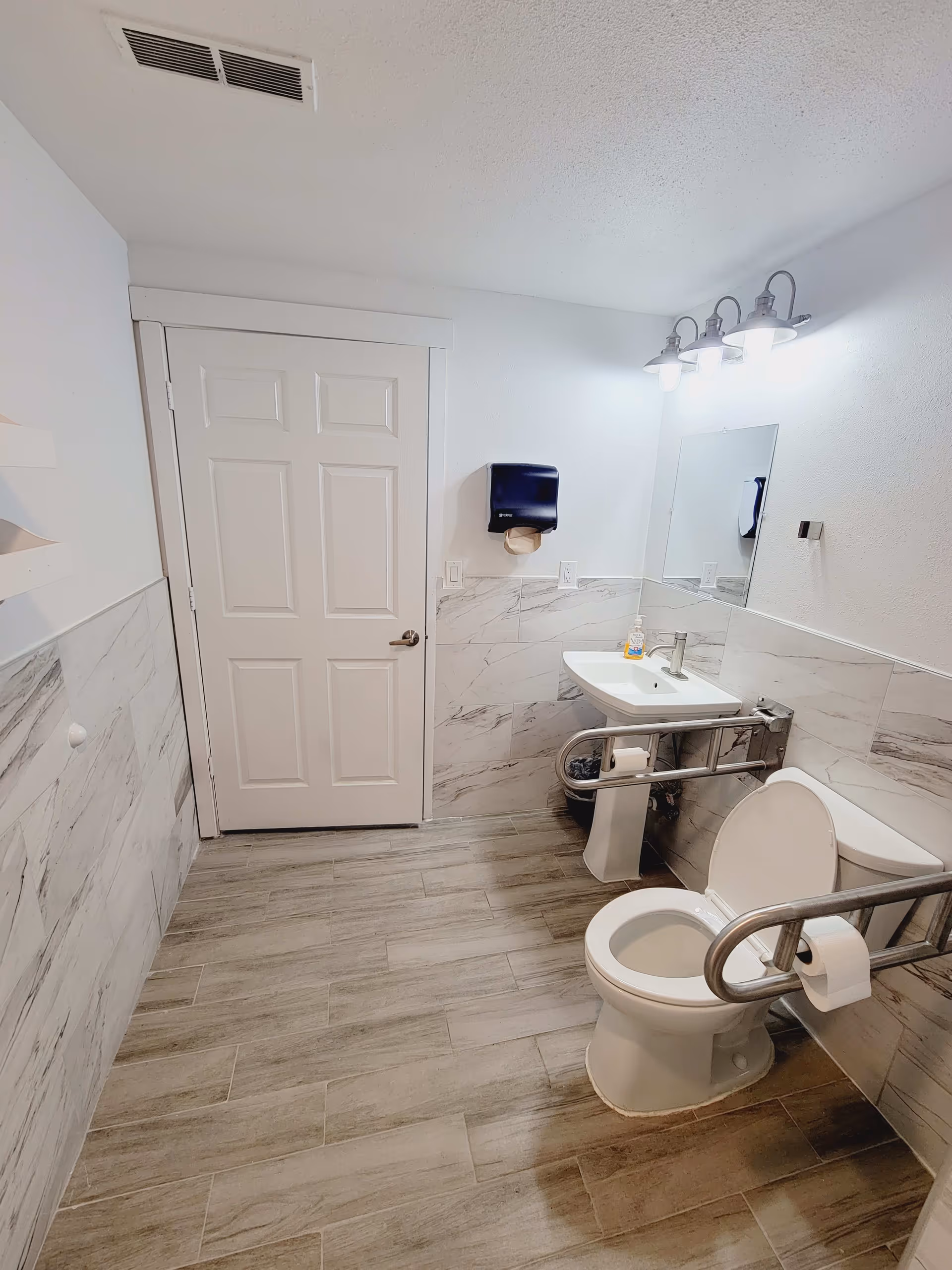 A clean, accessible bathroom with a white toilet equipped with metal grab bars on both sides, a white pedestal sink with a soap dispenser, a wall-mounted paper towel dispenser, a mirror above the sink, and a closed white door. The walls are partially tiled with a marble pattern, and the floor has wood-like tiles. There is a three-light fixture above the mirror providing bright lighting.
