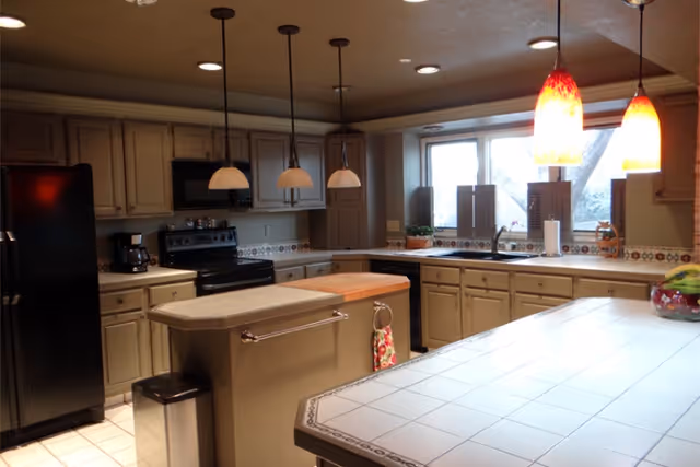 A spacious kitchen with beige cabinets, a black refrigerator, stove, and microwave. There are three pendant lights hanging over a central island with a towel hanging on the side. The kitchen has a large tiled countertop in the foreground and a window above the sink letting in natural light.