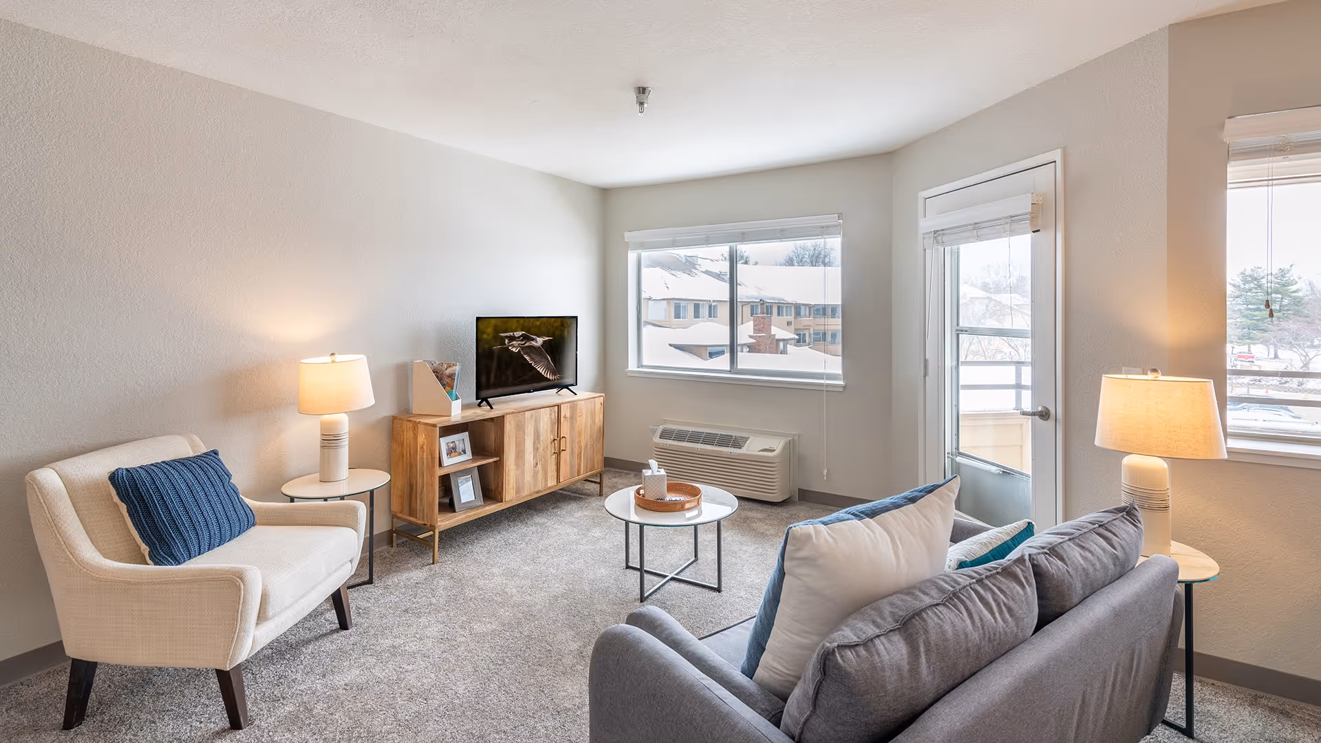 A bright and cozy living room with a gray sofa adorned with white and blue pillows, a beige armchair with a blue cushion, two side tables each with a lamp, a round coffee table, a wooden TV stand with a flat-screen TV, and large windows letting in natural light. There is a door leading to an outdoor balcony.