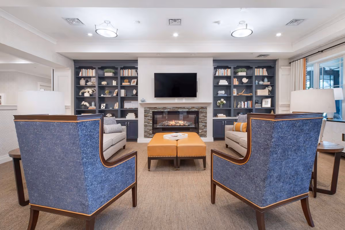Bright communal living room with two upholstered armchairs facing a fireplace and wall-mounted TV, sofas, ottoman and built-in bookshelves.