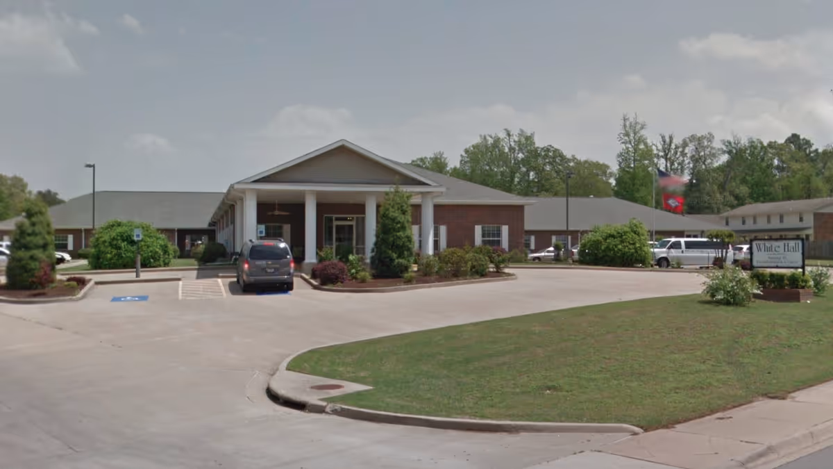 Exterior view of The Blossoms at White Hall Rehab & Nursing Center showing a single-story brick building with white columns at the entrance, a driveway with parked vehicles, landscaped bushes, and a sign near the road with the facility name.