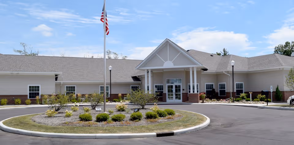 Front exterior view of Stone Creek Assisted Living and Memory Care building with a circular driveway, landscaped roundabout with bushes and small trees, an American flag on a flagpole, and a clear blue sky.