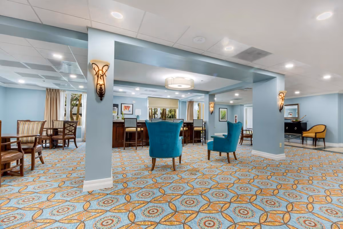Spacious, brightly lit communal lounge with patterned carpet, teal armchairs, tables and chairs under recessed lighting.