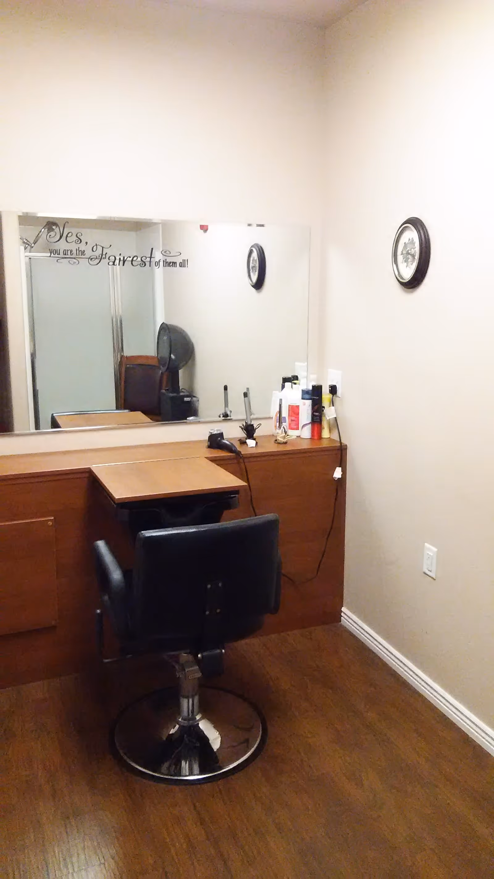 A small salon or grooming station with a black swivel chair in front of a wooden counter. On the counter are various hair care products, a hairdryer, and curling irons. A large mirror is mounted on the wall behind the counter, reflecting part of the room including a shower stall and a clock on the opposite wall. The wall above the mirror has a decorative quote that reads, 'Yes, you are the Fairest of them all!'
