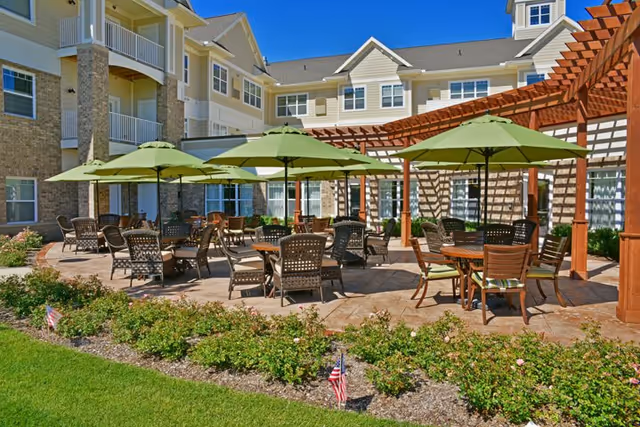 Courtyard patio with wicker tables and chairs under green umbrellas and a wooden pergola outside a multi-story senior living building.