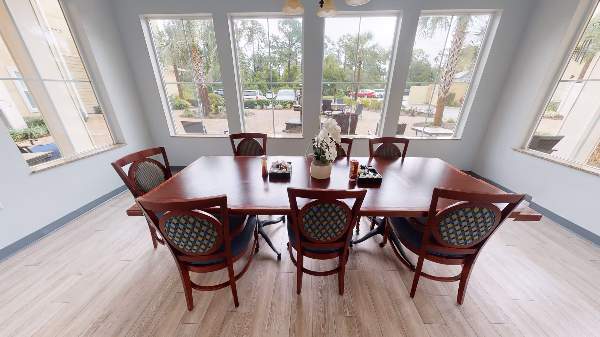 A dining area with a rectangular wooden table surrounded by six wooden chairs with patterned cushions. The room has large windows providing a view of an outdoor patio area with palm trees and parked cars. The table has a small flower arrangement and condiment holders.
