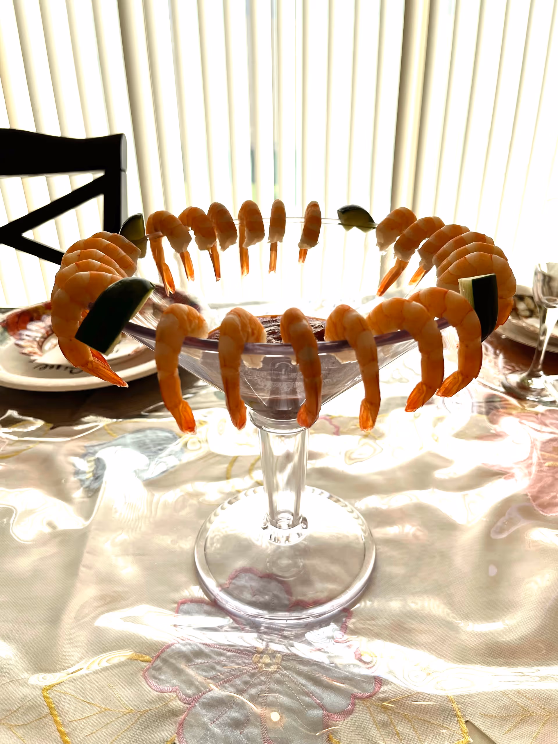 A clear glass shrimp cocktail dish filled with cocktail sauce and garnished with cooked shrimp and lime wedges, placed on a table with a floral tablecloth and a chair in the background.