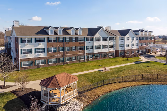 Three-story senior living building beside a pond with a gazebo, walking path, and clear blue sky.