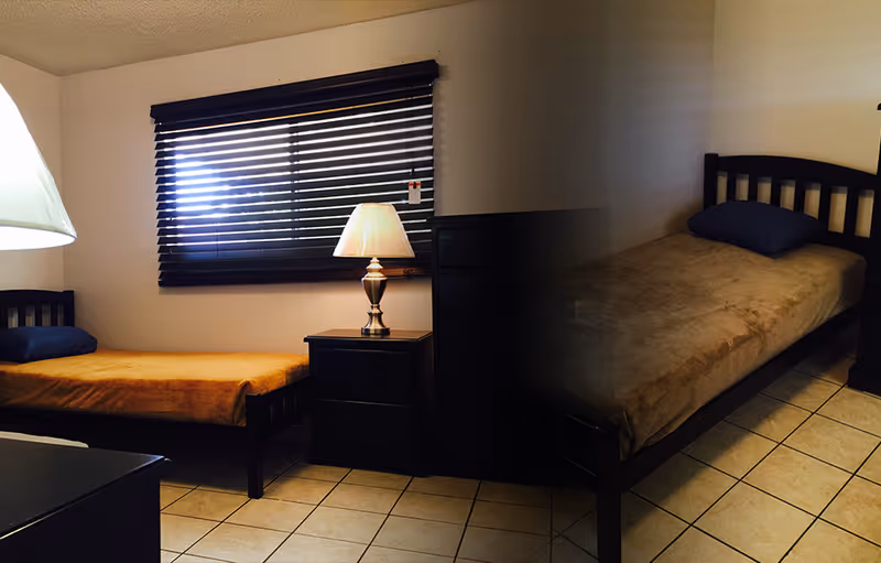 A small bedroom with two single beds, each with a pillow and a brown blanket. Between the beds is a dark wooden nightstand with a lamp on it. The room has tiled flooring and a window with dark blinds partially open.