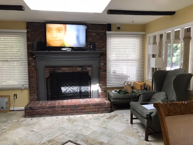 A cozy living room area with a brick fireplace and a mounted flat-screen TV above it displaying a close-up of a child's face. The room has two windows with white blinds on either side of the fireplace. There is a green upholstered armchair and an ottoman with some items on it, a wooden chair partially visible in the foreground, and a lamp near the window. The floor is tiled with a light-colored pattern.