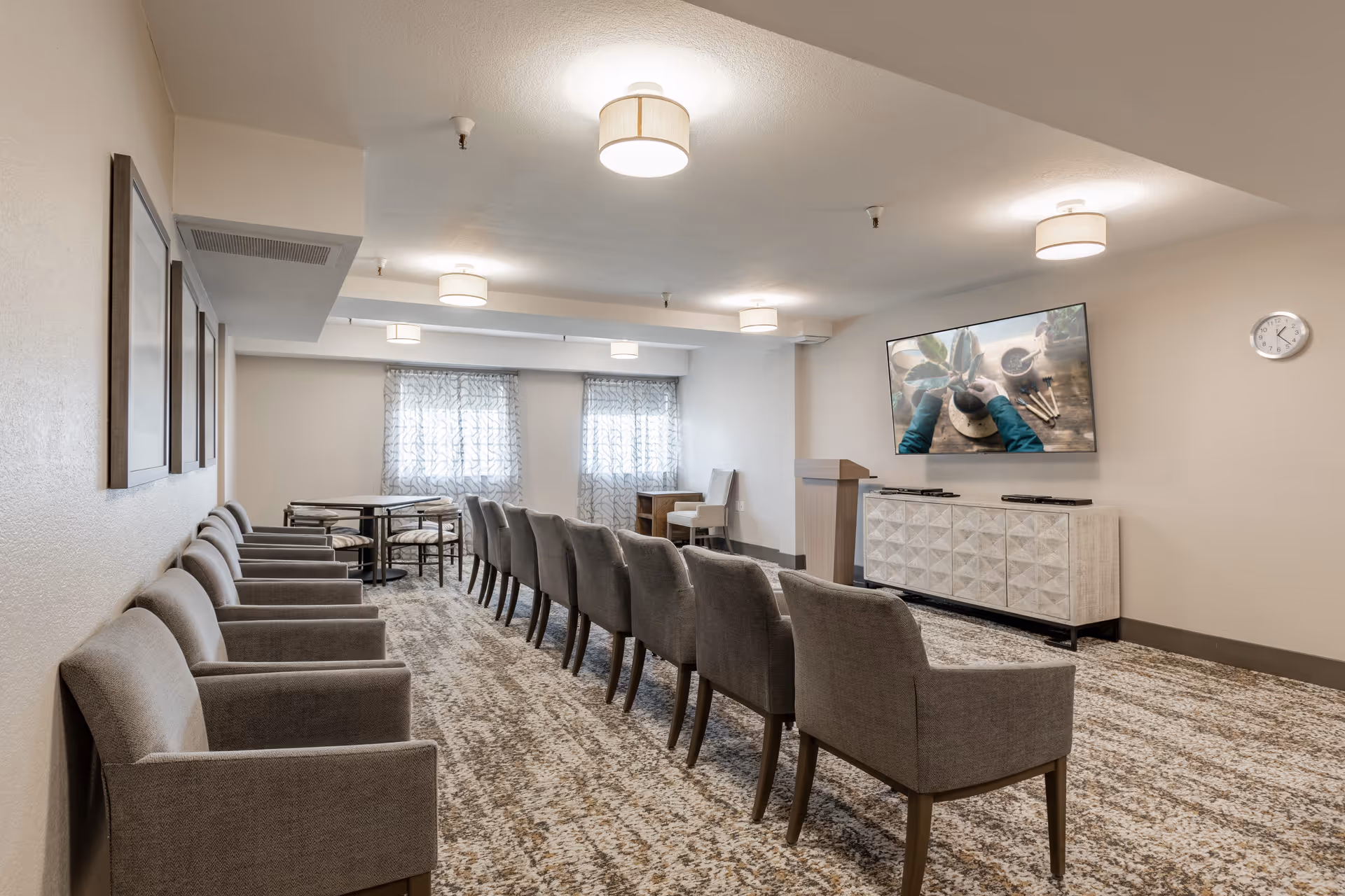 A meeting or presentation room with two rows of gray upholstered chairs facing a large flat-screen TV mounted on the wall. The TV displays an image of hands working with plants. The room has patterned carpet, light-colored walls, ceiling lights, two windows with sheer curtains, a clock on the wall, and a white cabinet beneath the TV.