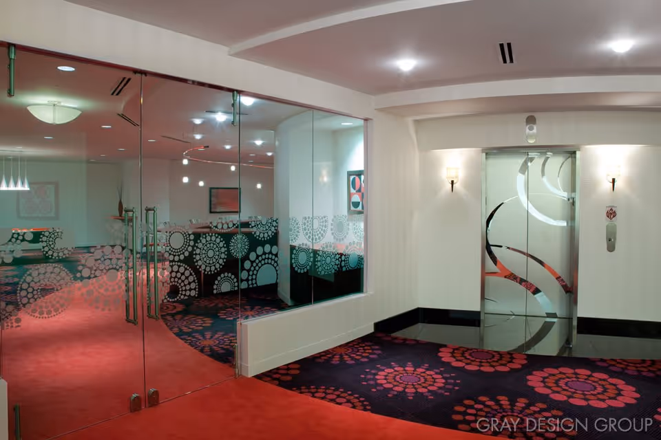 Interior view of a modern hallway with patterned carpet and glass doors leading to a conference or meeting room. The hallway features an elevator with decorative glass doors and wall sconces providing lighting.