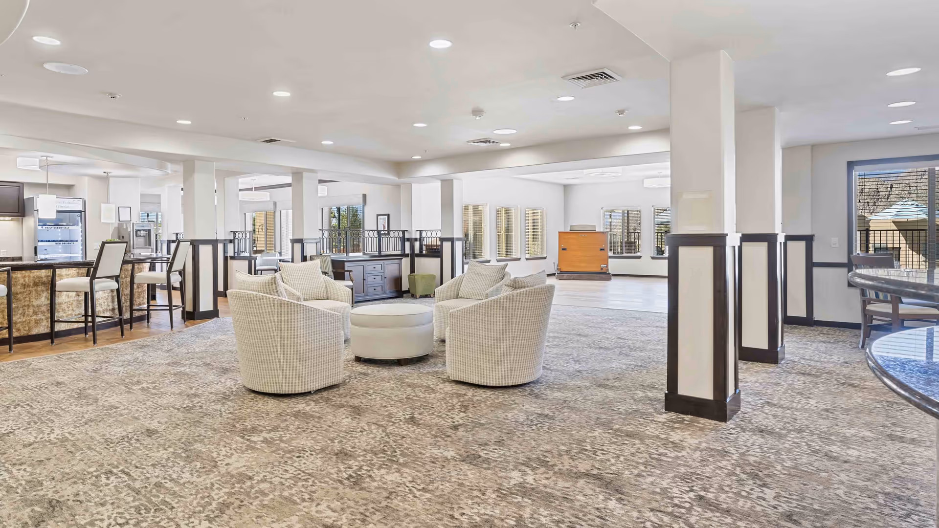 Spacious and well-lit common area in a retirement living facility featuring a seating arrangement with four cushioned chairs around a round ottoman, a carpeted floor, and a bar area with high chairs in the background. Large windows allow natural light to fill the room.