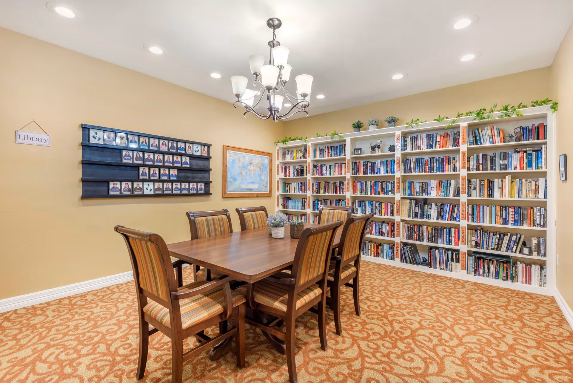 A bright library room with a central wooden table and chairs, bookshelves lining the right wall, and a 'Library' sign on the left wall.