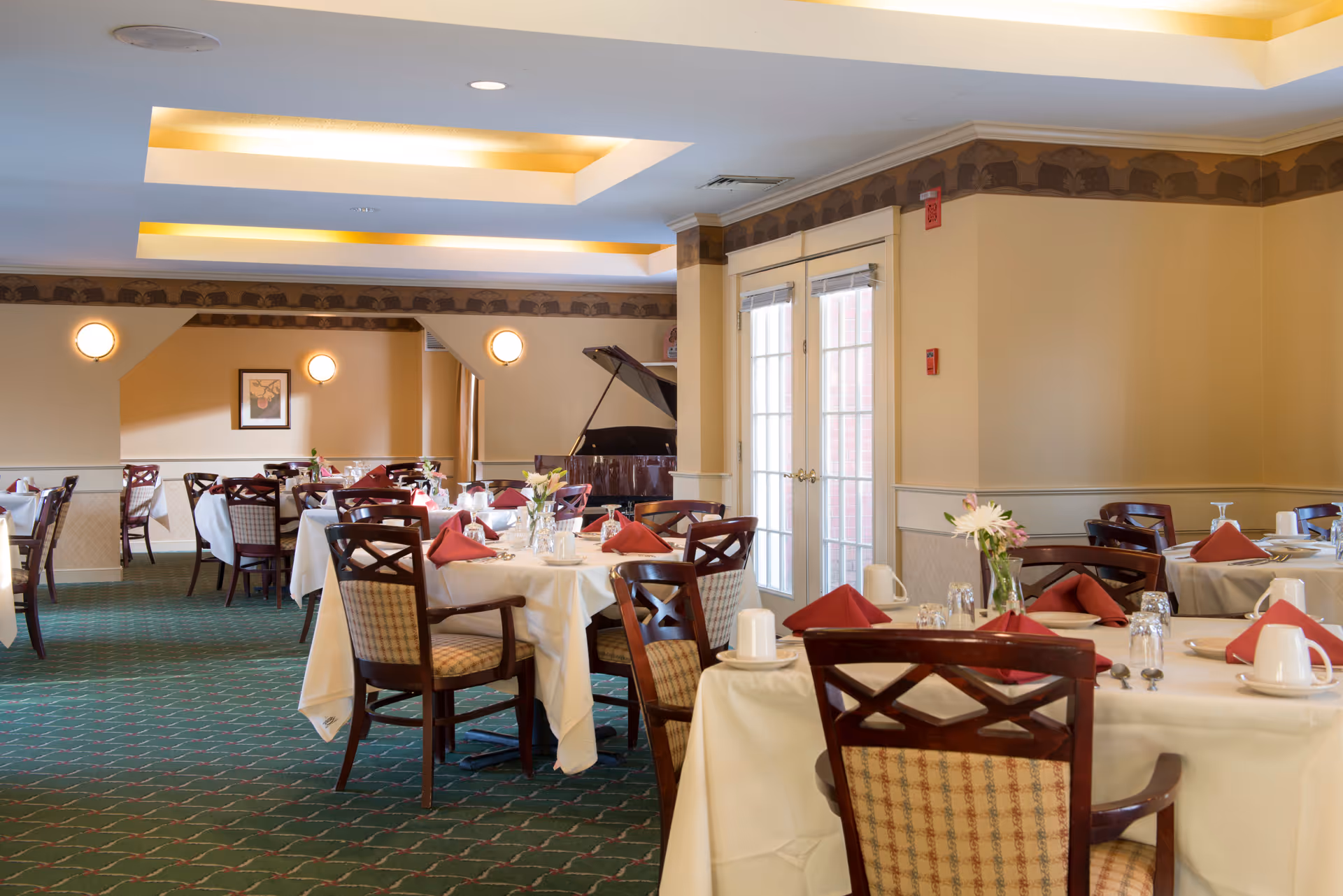 A dining room with several tables covered in white tablecloths, each set with red folded napkins, cups, glasses, and silverware. The room has green carpet with a pattern, beige walls with a decorative border near the ceiling, and a grand piano in the corner near double glass doors.