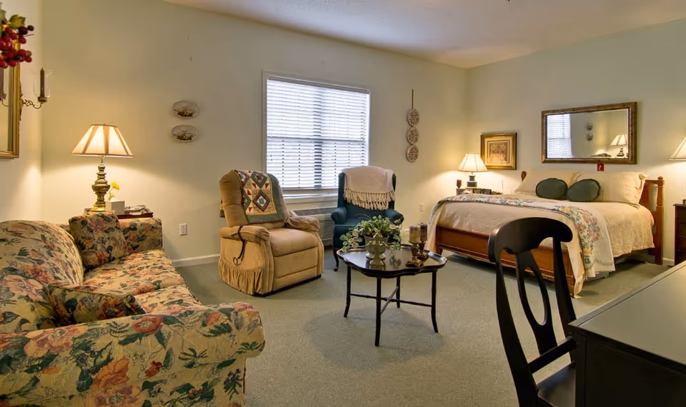 A cozy senior living bedroom with a floral patterned sofa, two armchairs, a wooden bed with a quilted bedspread, a coffee table with decorative items, a desk with a chair, and several lamps providing warm lighting. The room has a window with blinds and light-colored walls.