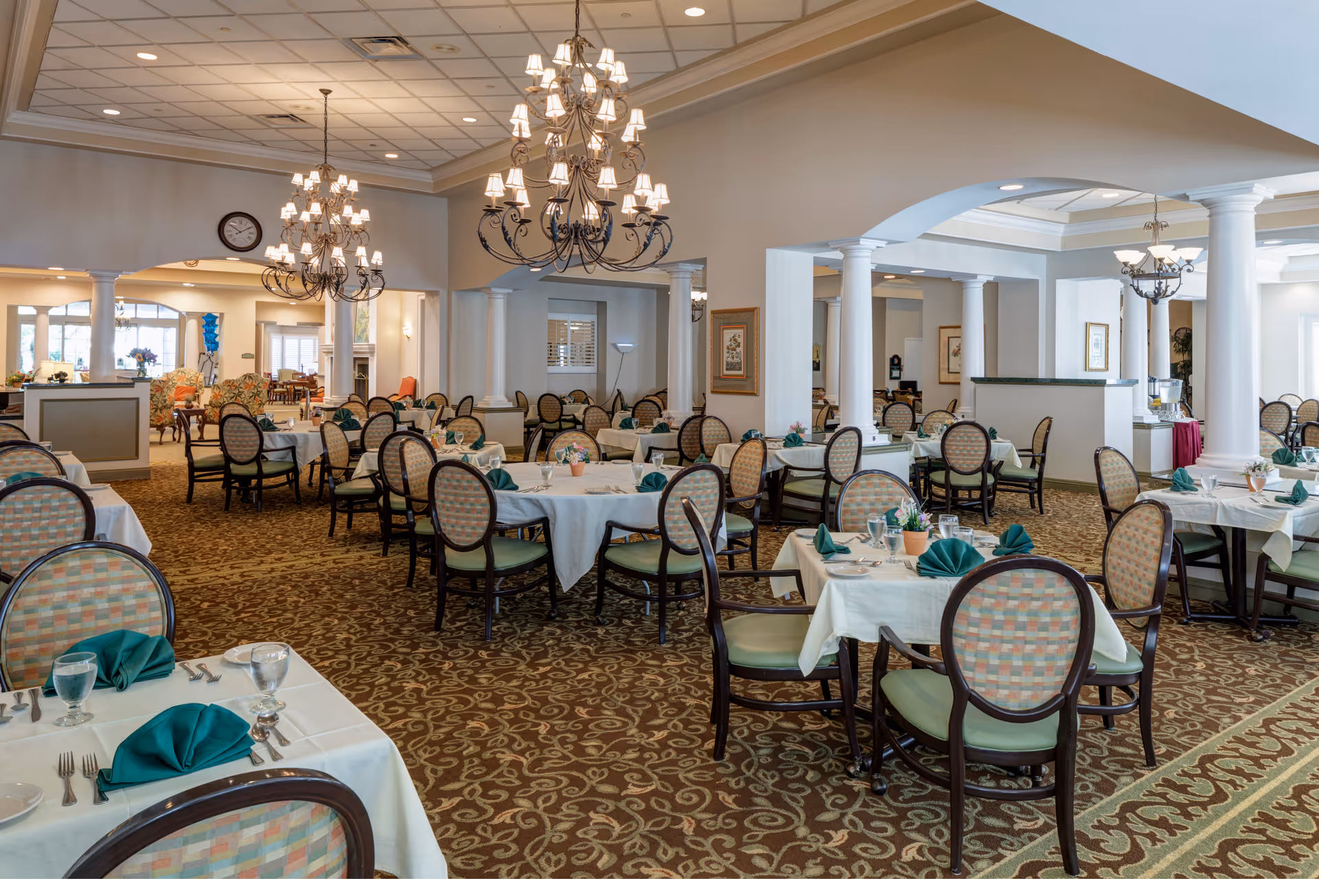 A spacious dining room with multiple round tables covered with white tablecloths, each set with green folded napkins, glassware, and silverware. The room features patterned carpet, elegant chandeliers hanging from the ceiling, and decorative columns. There are upholstered chairs around the tables and a clock on the wall in the background.