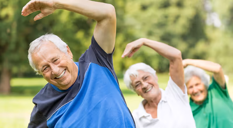 Three elderly individuals outdoors stretching their arms overhead and smiling, with greenery in the background.