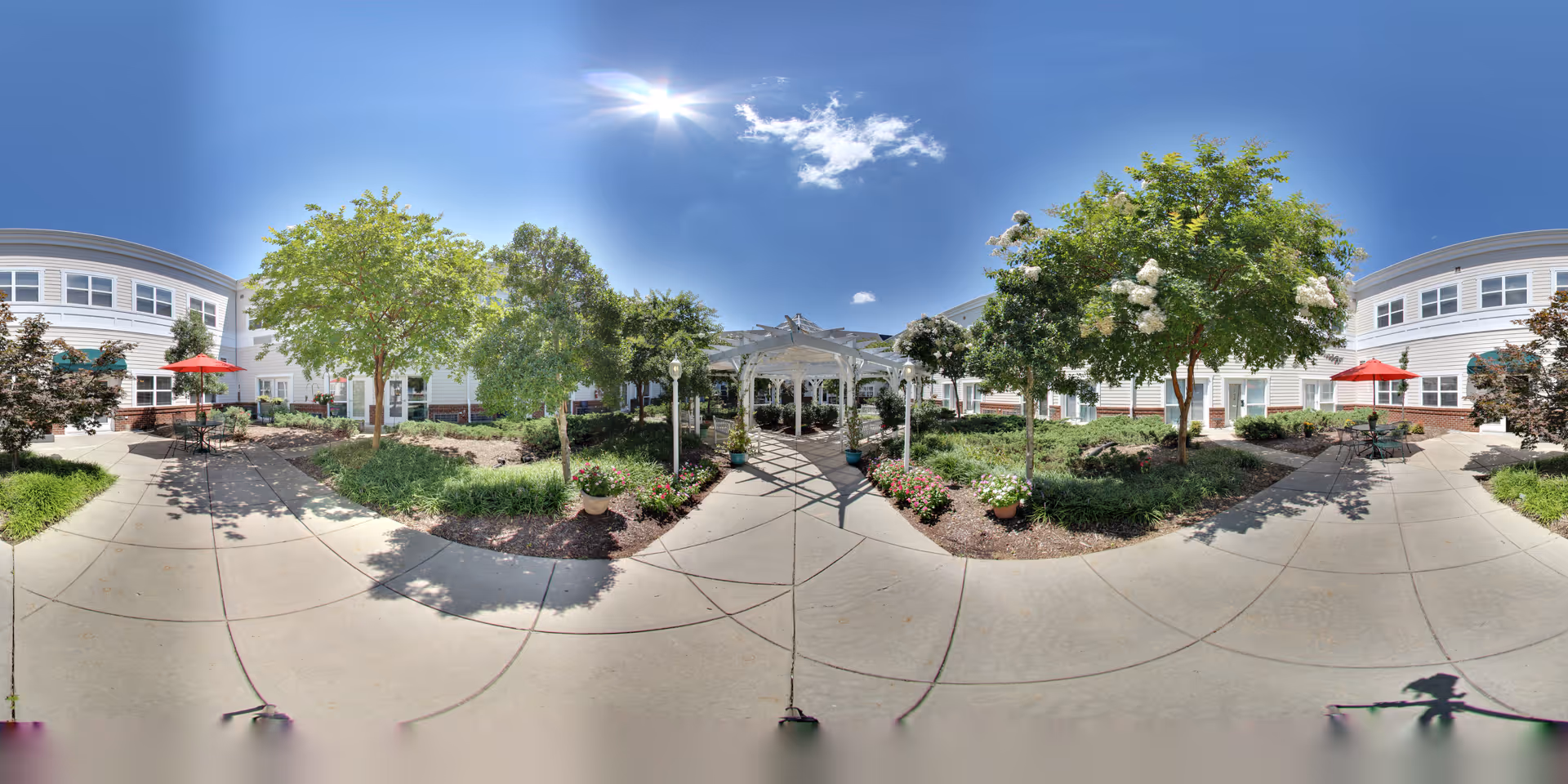 Outdoor courtyard area at Morningview at Irving Park featuring a paved walkway, green trees, flowering plants, and a white pergola in the center. The building surrounds the courtyard with windows and red umbrellas shading outdoor tables.