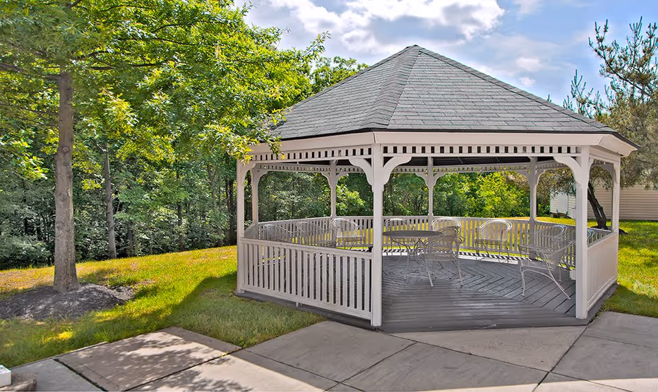 A white wooden gazebo with a gray shingled roof situated on a concrete patio surrounded by green grass and trees under a partly cloudy sky.