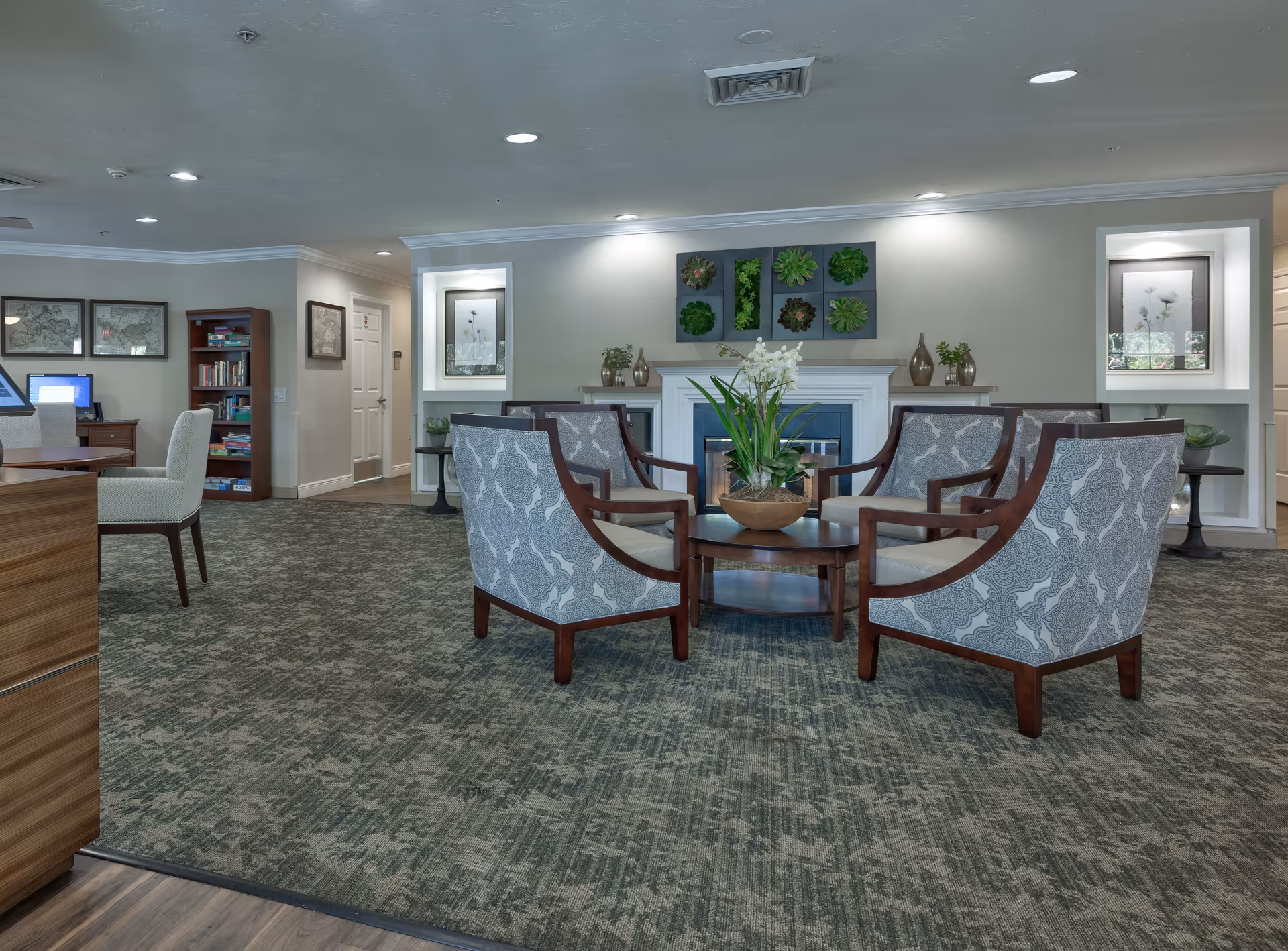 A cozy common area in a senior living facility featuring a circular arrangement of four patterned armchairs around a wooden coffee table with a potted plant centerpiece. The room has a carpeted floor, a white fireplace with decorative plants on the mantel, framed artwork on the walls, and built-in shelves with books and decor. The lighting is soft with recessed ceiling lights.