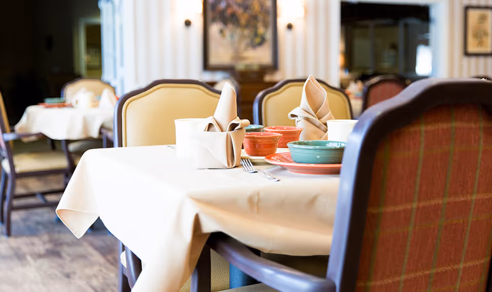 A dining table set with beige tablecloth, folded beige napkins, colorful bowls and plates, and silverware in a well-lit dining room with upholstered chairs and framed artwork on the wall.
