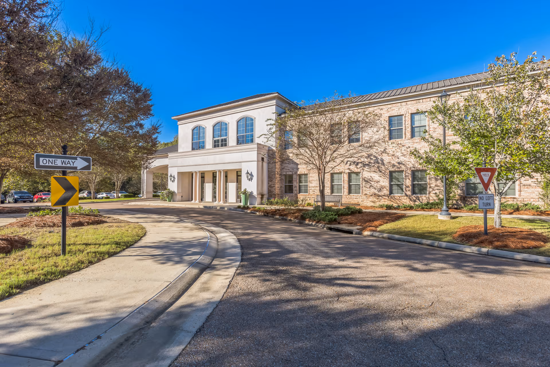 Exterior view of a two-story senior living facility building with a covered entrance, surrounded by trees and landscaping under a clear blue sky. There are traffic signs indicating one way and no left turn near the driveway.