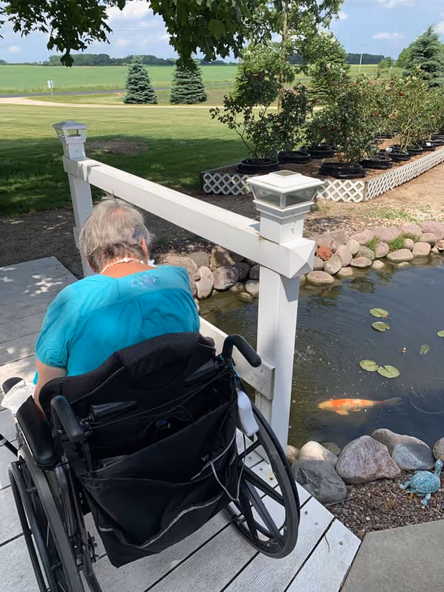 An elderly person in a wheelchair wearing a turquoise shirt is sitting on a wooden deck next to a small pond with koi fish and lily pads. The deck has white railings with lantern-style lights. In the background, there is a garden area with potted plants and a grassy field with trees under a partly cloudy sky.