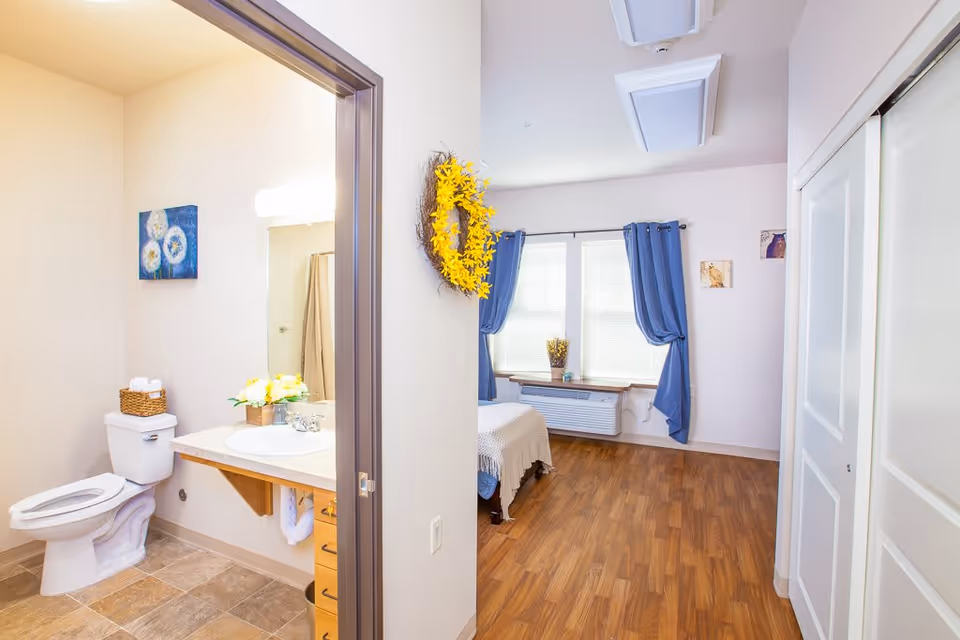 View of a senior living facility room with a bathroom on the left featuring a toilet, sink, and a basket with towels. The main room has wooden flooring, a bed with a white blanket, blue curtains on the windows, a yellow wreath on the wall, and some small wall decorations.