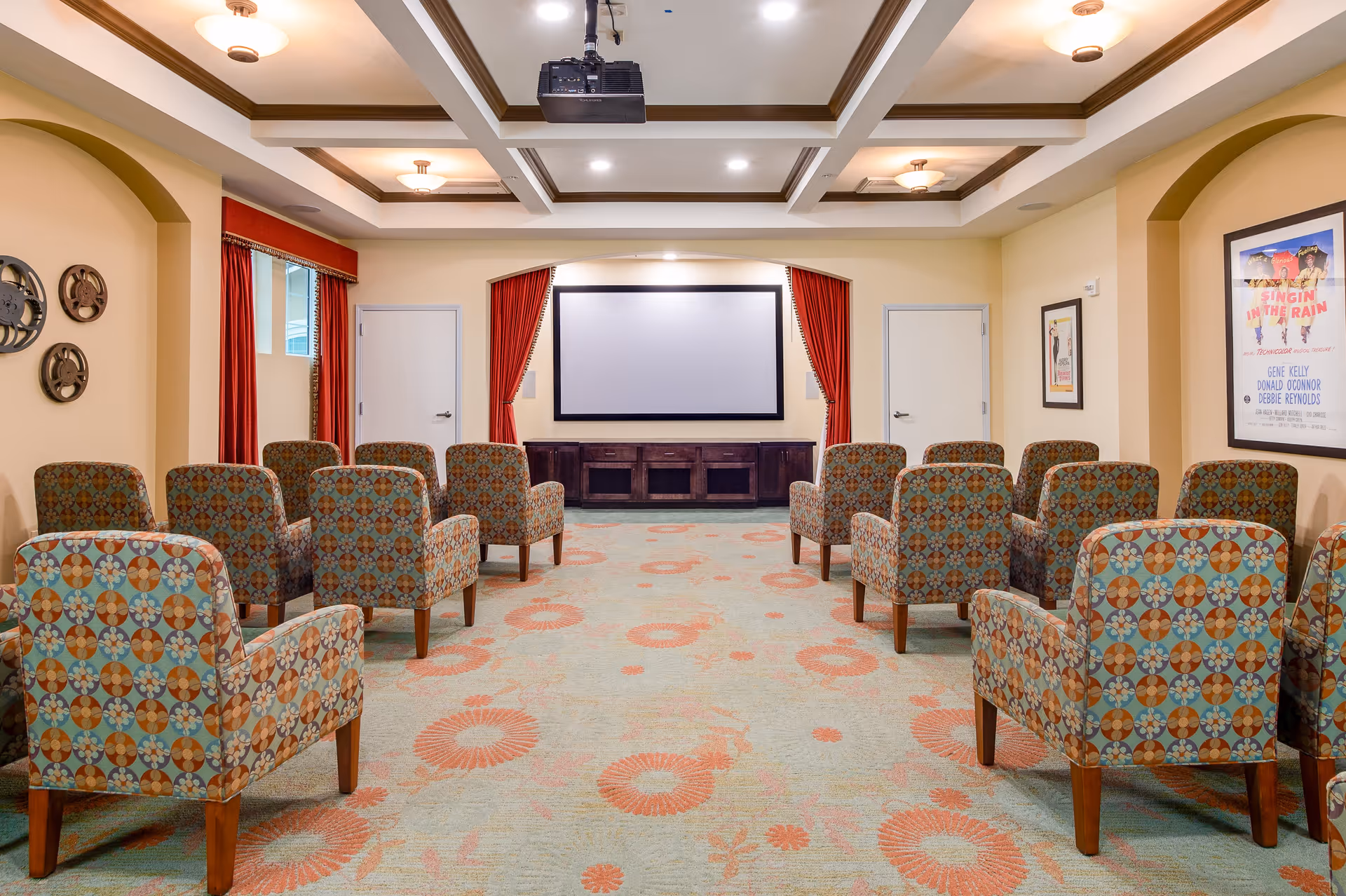 A small theater room with patterned upholstered chairs arranged in rows facing a large projection screen. The room has a coffered ceiling with recessed lighting and wall sconces, red curtains framing the screen, and movie posters on the walls.