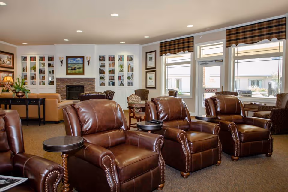 A cozy living room area in Whitetail Springs Memory Care Community featuring four brown leather recliners with small round side tables, a fireplace with a stone surround and built-in white bookshelves filled with books and decorative items, large windows with plaid valances, and additional seating including armchairs and a sofa.