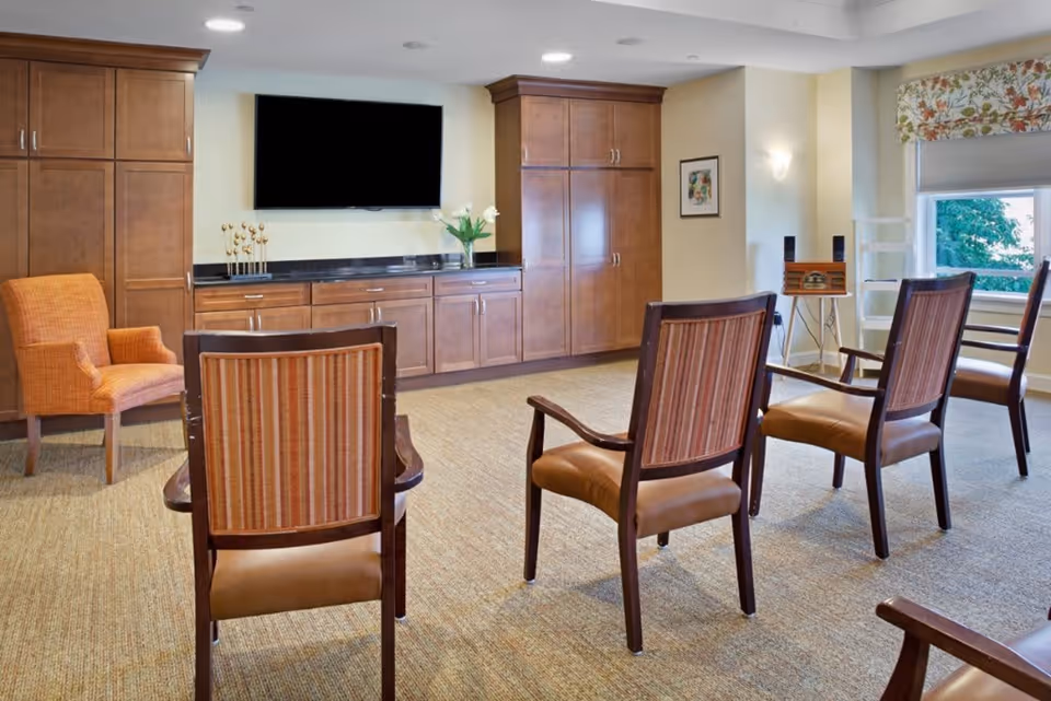 A well-lit common area with several wooden chairs arranged facing a wall-mounted flat screen TV. The room features wooden cabinetry with a black countertop, a vase with flowers, an orange armchair, a window with floral valance, and a small vintage-style radio on a stand.