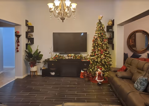 A cozy living room decorated for Christmas with a lit Christmas tree adorned with ornaments and lights on the right side. A brown leather sofa with pillows is placed next to the tree. A flat-screen TV is mounted on the wall above a black entertainment unit decorated with garlands and Christmas ornaments. Shelves with decorative items flank the TV. A chandelier hangs from the ceiling, and a round mirror is visible on the right wall.