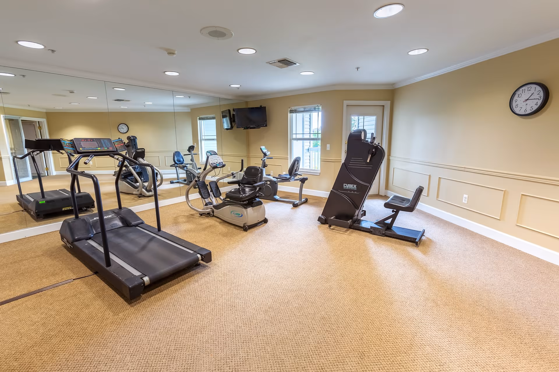 A fitness room with exercise equipment including a treadmill, stationary bikes, and a Cybex exercise machine. The room has beige walls, a large mirror on one side, a wall clock, two windows, and a wall-mounted TV.