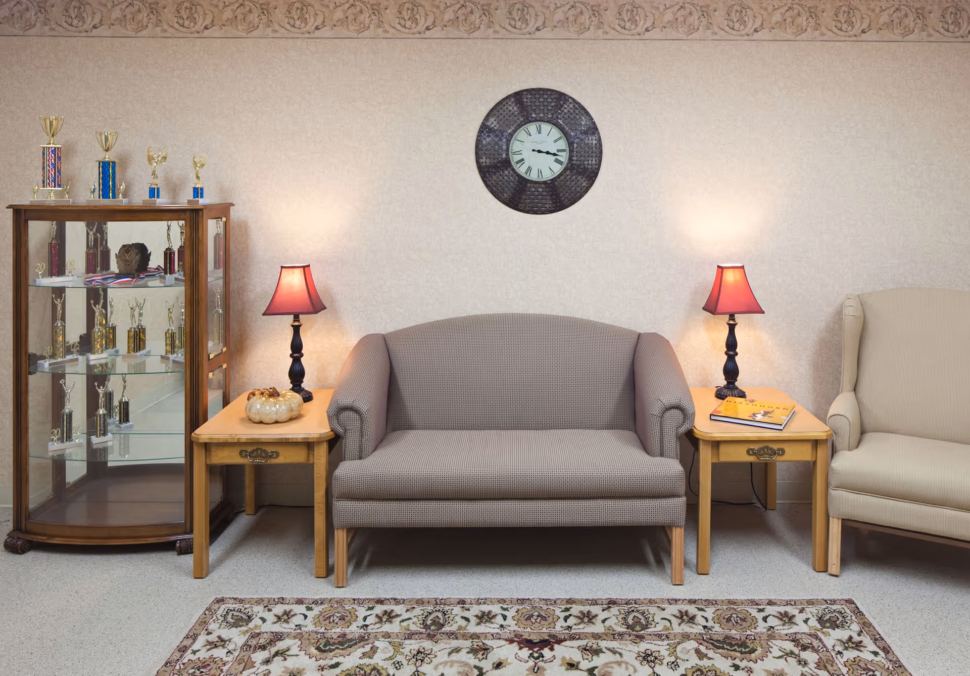 A cozy sitting area with a patterned loveseat centered between two wooden side tables, each holding a lamp with a red shade. To the left, there is a glass display cabinet filled with trophies and awards. On the wall above the loveseat, there is a round clock. To the right of the loveseat, there is a beige upholstered armchair. A decorative rug with floral patterns covers the floor in front of the seating.