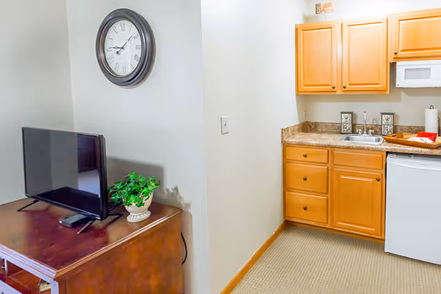 Interior view showing a small kitchenette with wooden cabinets, a countertop with a sink, a microwave, and a mini refrigerator. Adjacent to the kitchenette is a wooden table with a flat-screen TV and a small potted plant. A wall clock is mounted above the table on a light-colored wall.