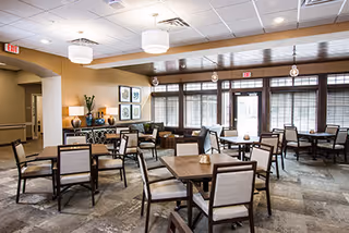 Bright, well-lit communal dining room with multiple square tables and chairs, a sideboard with lamps, and large windows along one wall.