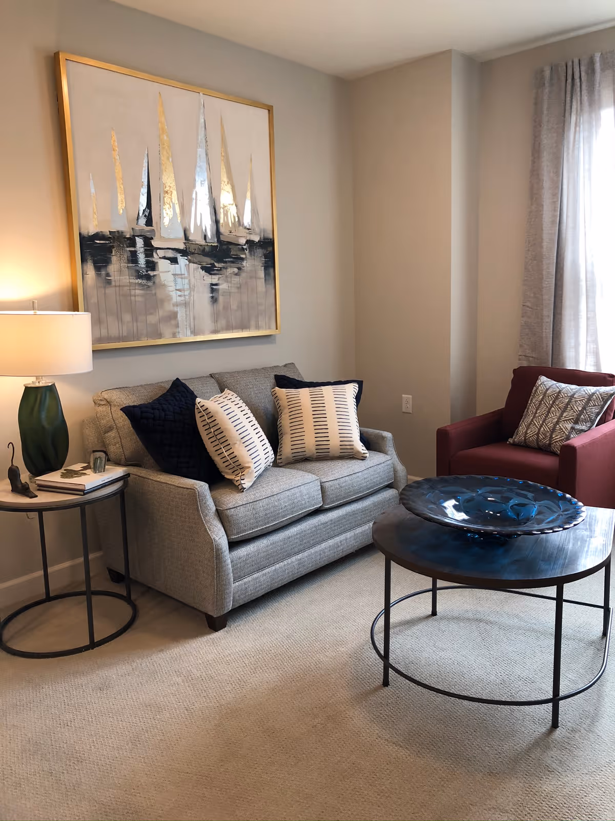 A cozy living room featuring a gray loveseat with pillows, a red armchair, round coffee and side tables, and sailboat wall art.